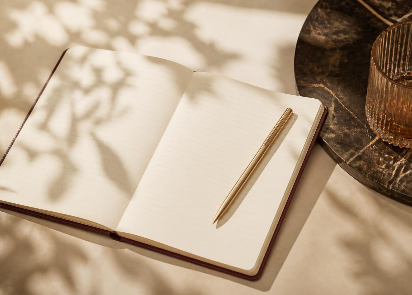 Open ivory notebook with a gold pen beside a marble tray and amber glass